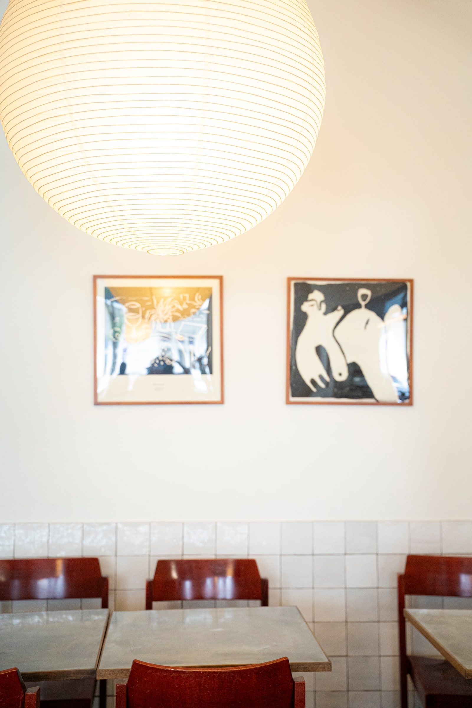 Zwei gerahmte Drucke an einer cremefarbenen Wand über roten Stühlen, unter einer Papierlampe